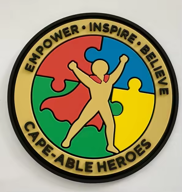 Round Cape-Able Heroes PVC patch: a tan hero with a red cape, arms raised, over red, blue, green, and yellow puzzle pieces, ringed with Empower, Inspire, Believe and Cape-Able Heroes
